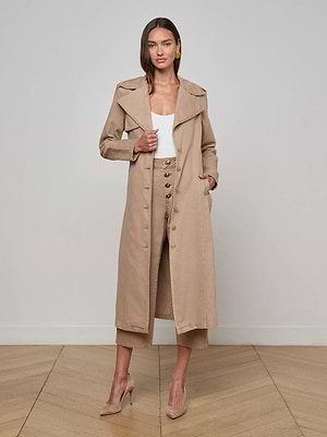 A woman stands indoors on a wood floor wearing the L'AGENCE Zavia Jean Trench Coat over a white top and tan high-waisted pants with large buttons, paired with tan heels. She poses with one hand in her pocket.
