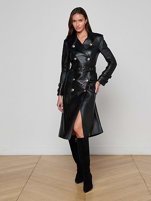 A woman stands indoors on a wooden floor, confidently posing against a light wall in the L'AGENCE Celina Leather Trench Coat, featuring gold buttons and a front slit, paired with knee-high black suede boots.