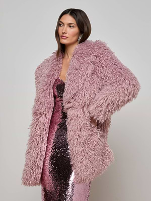 A woman models the L'AGENCE Toira Faux Fur Coat, a long plush curly-texture light pink piece, over a shimmering pink sequin dress against a plain light gray background.