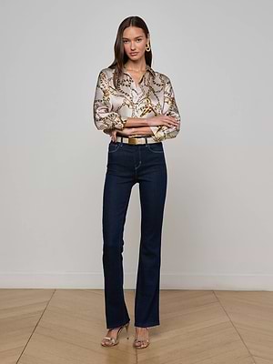 A woman with long brown hair poses confidently indoors, wearing L'AGENCE Ruth Straight-Leg Jeans, a patterned chain-print blouse, gold belt, hoop earrings, and heeled sandals against a plain wall.