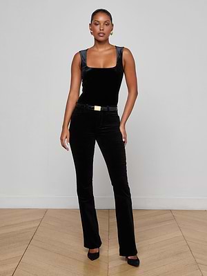 A woman with short hair and a confident expression stands against a light gray wall, wearing L'AGENCE Stevie Velvet Jeans and a matching sleeveless black velvet top, styled with a black belt featuring a gold buckle and black heels.