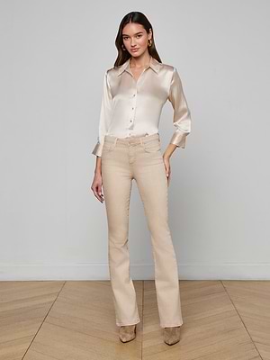A woman with long hair and gold hoop earrings stands in a minimalist room wearing a shiny beige satin blouse, the Selma Bootcut Jean with a contoured waistband, tan heeled boots, and maintains a neutral expression, arms relaxed by her sides.