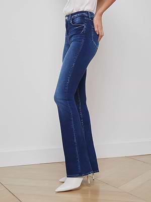 A person wearing L'AGENCE Selma Bootcut Jeans in dark blue stretch denim, a white top, and white pointed-toe high heels stands against a white wall on a light wooden floor.