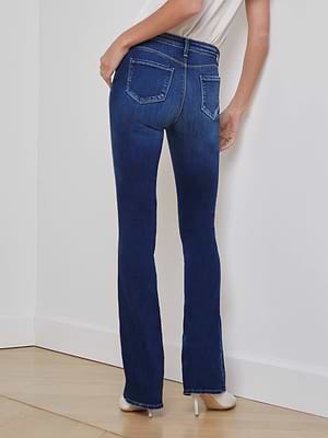 A person stands with their back to the camera against a white wall and light wood floor, wearing L'AGENCE's Selma Bootcut Jean in dark blue stretch denim and white high-heeled shoes.