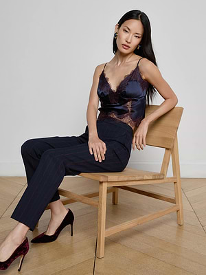 A woman with long dark hair sits on a wooden chair, wearing L'AGENCE Rebel Pinstriped Trouser, a navy lace-trimmed camisole, and burgundy heels, against a plain light-colored wall and wooden floor.