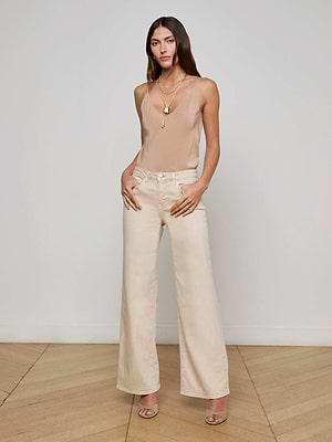 A woman with long brown hair wears the L'AGENCE Alicent Wide-Leg Jean, a beige tank top, and heeled sandals. She stands indoors on a wooden floor with layered gold necklaces and a neutral expression against a plain white wall.