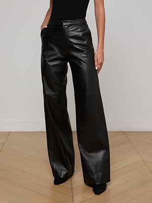 A person wearing L'AGENCE Livvy Leather Trouser, crafted from premium lambskin in a wide-leg black silhouette, stands with hands in pockets on a light wood floor against a plain white wall; only the lower body is shown.