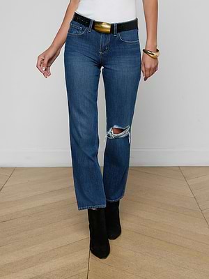 A person in Nevia Slouchy Straight-Leg Jeans with a ripped knee, a black belt, white top, gold bracelet, and black heeled boots stands on a wooden floor against a plain white wall.