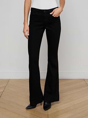 A person models the L'AGENCE Lenora Low-Rise Flare Jean in black wash with black pointed-toe shoes, standing on a wooden floor against a plain white wall, showing only their lower torso and legs.