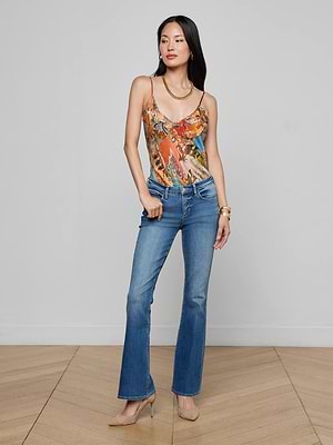 A woman with long dark hair poses indoors against a plain light wall, wearing the L'AGENCE Cara Low-Rise Bootcut Jean in stretch denim, a colorful sleeveless top, beige heels, and gold jewelry. One hand rests in her pocket on a wooden floor.