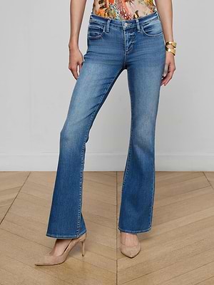 A person wears L'AGENCE Cara Low-Rise Bootcut Jeans in blue stretch denim, paired with a colorful top and beige pointed-toe heels. Only their lower torso and legs are visible, standing on a wooden floor against a white wall.
