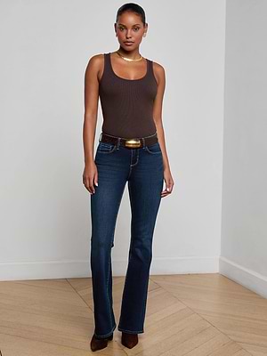 A woman stands indoors wearing L'AGENCE Cara Low-Rise Bootcut Jeans in dark blue super stretch denim, paired with a brown tank top, black belt, brown pointed shoes, and gold jewelry. She poses against a plain white wall on light wood flooring.
