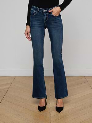 Wearing Cara Low-Rise Bootcut Jeans in dark blue, a black long-sleeve top, and black pointed-toe heels, this individual stands on a wooden floor against a plain white wall.