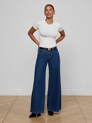 A woman poses confidently against a white wall, wearing a fitted white short-sleeve top and L’AGENCE Savina Low-Rise Wide-Leg Jeans in indigo wash, paired with a black belt and shoes. Her hands are behind her back as she looks at the camera.