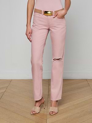A person stands on a wooden floor wearing the Armida Low-Rise Slouch Jean in light pink with a ripped knee, paired with a beige belt featuring a shiny gold buckle and beige high-heeled sandals. The background is a plain white wall.