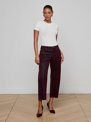 A person stands indoors against a plain wall, wearing a fitted white short-sleeve top, L'AGENCE Dolores Flocked Bow-Leg Jean in bordeaux velvet, and matching burgundy heels on a light wooden floor.