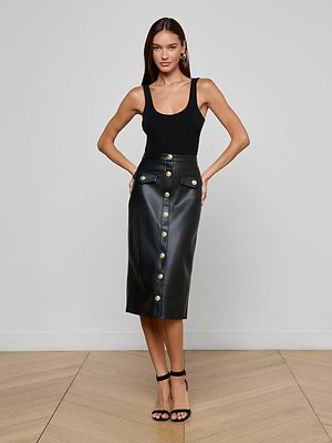 A woman stands confidently in a minimalist room, wearing a sleeveless black top, black heels, and the L’AGENCE Milann Vegan Leather Skirt—a high-waisted pencil skirt with gold accent buttons. Her hands rest on her hips as she faces the camera.
