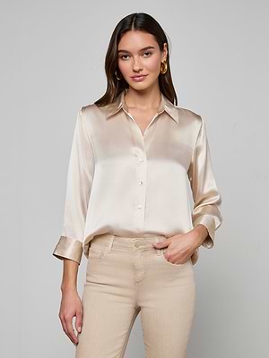 A woman in the slim-fit Dani Silk Blouse, a champagne-colored long-sleeve top, paired with beige pants, stands against a plain background with a slight smile and one hand in her pocket.