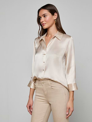 A woman with long brown hair wears the Dani Silk Blouse, a slim-fit champagne top with rolled sleeves, paired with beige pants and standing against a plain light gray background.