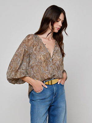 A woman with long brown hair wears the Teagan Blouse in paisley crinkle poly chiffon with billowy sleeves, paired with blue jeans and a wide gold belt, standing slightly to the side against a plain background.