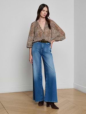 A woman poses indoors with one hand in her pocket, wearing the Teagan Blouse—a semi-sheer paisley print top with billowy sleeves—paired with wide-leg blue jeans, a brown belt, and brown pointed-toe boots against a plain white wall.