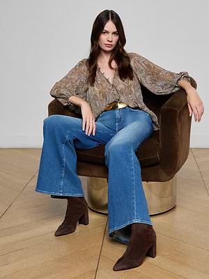 A woman with long brown hair sits in a brown swivel chair, wearing the Teagan Blouse with blue flared jeans, a gold belt, and brown heeled boots, her arm resting on the chair’s back.
