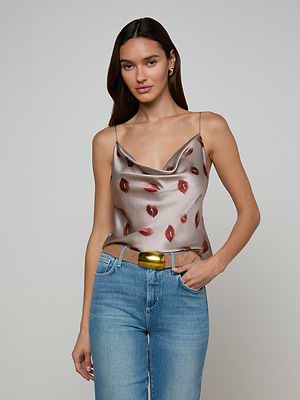 A woman with long brown hair wears the Calista Satin Camisole—silver with a red lip print and adjustable straps—paired with blue jeans and a gold belt, standing against a plain light gray background.