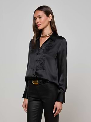 A woman poses against a plain backdrop, wearing the L'AGENCE Divya Silk Shirt in black with pleated details and satin buttons, paired with black pants, gold hoop earrings, layered gold necklaces, and a black belt with a gold buckle.