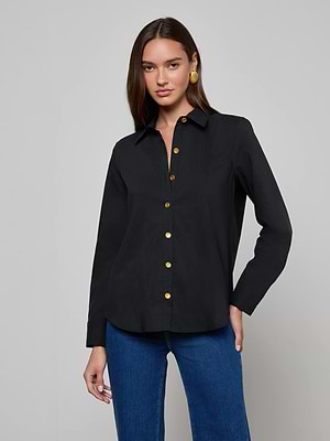 A woman wears the L'AGENCE Ellery Button-Down Shirt—black, long-sleeve, relaxed fit with gold buttons—paired with blue jeans. She has long brown hair, gold earrings, and stands against a plain light gray background.