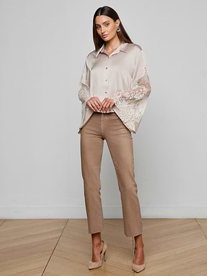 A woman poses indoors on a wooden floor against a light gray wall, wearing the Capri Lace Sleeve Blouse—beige with lace bell sleeves—paired with tan pants and nude heels. She has straight brown hair.