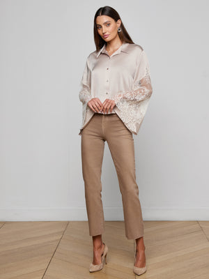 A woman poses indoors on a wooden floor against a light gray wall, wearing the Capri Lace Sleeve Blouse—beige with lace bell sleeves—paired with tan pants and nude heels. She has straight brown hair.