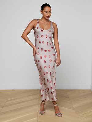 A woman poses against a white wall on a wooden floor, wearing the Akiya Satin Maxi Dress—a light pink, bias-cut evening gown with red lip prints—paired with strappy silver heels and delicate jewelry.
