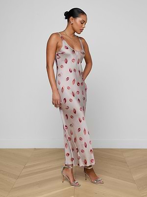 A woman stands sideways in the Akiya Satin Maxi Dress, a long, sleeveless satin evening gown with a pink base and red lip print. Her hair is pulled back, and she pairs it with silver high-heeled sandals and delicate necklaces.