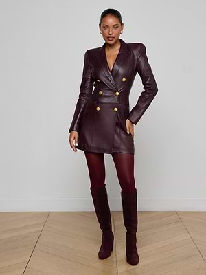A woman stands indoors against a plain wall, wearing the Tora Leather Blazer Dress in burgundy with gold buttons, matching tights, and knee-high suede boots. Her hair is pulled back and one hand rests on her hip.