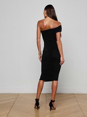 A woman with long brown hair faces away, wearing the Cheryl Shirred Midi Dress for a flattering fit, paired with black high-heeled sandals, standing against a plain light-colored wall and wooden floor.