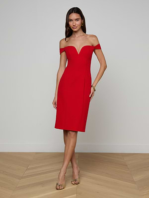A woman poses indoors against a white wall wearing the Vivie Dress—a knee-length, fitted red dress with off-the-shoulder straps and a sculpted corset bodice. She styles it with long hair, hoop earrings, a bracelet, and open-toe heels.