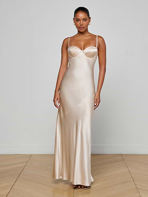 A woman stands indoors on a wooden floor, wearing the Sayla Silk Gown by L'AGENCE—a long champagne gown with thin straps and a fitted bodice. She exudes vintage glamour, facing the camera with arms relaxed by her sides against a plain light background.