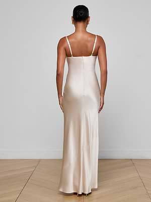 A woman, seen from behind in a simple room with light walls and a wooden floor, wears the Sayla Silk Gown by L’AGENCE—a long champagne dress with thin straps.