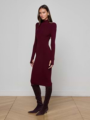 A woman poses on a light wood chevron floor against a plain gray wall, wearing the L'AGENCE Larelle Midi Dress—a ribbed knit, long-sleeve maroon dress—with matching knee-high boots, one foot slightly forward.