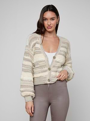 A woman with long dark hair wears the Harriet Cardigan, a cozy wool-blend knit with blouson sleeves, layered over a white top and taupe pants, standing against a plain light background.