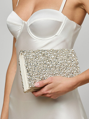 A woman in a white satin dress holds the Verve Crystal Clutch, adorned with handcrafted crystals, making it the perfect statement accessory at her side.
