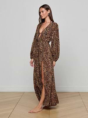 A woman stands barefoot on a wooden floor, wearing the L'AGENCE Sara Maxi Cover-Up Dress—a long, flowy leopard print cover-up with long sleeves, deep V-neckline, belted waist, and high slit—facing slightly left against a plain wall.