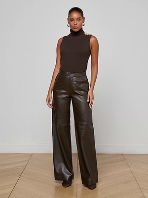 A woman stands indoors against a plain wall, wearing a sleeveless brown turtleneck top with gold buttons and high-waisted Livvy Leather Trousers by L'AGENCE. Her hair is pulled back and she pairs the look with heels.