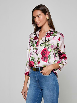 A woman with long brown hair wears the Dani Silk Blouse—a white, button-down top with a red rose print—paired with blue jeans, statement gold earrings, and a black belt. She stands against a plain light gray background, looking to the side.