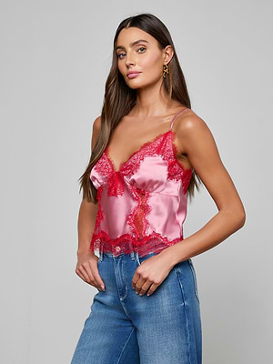 A woman with long brown hair models the Kimber Silk Lace Camisole, a cropped satin top with red lace trim, paired with blue jeans. She stands angled slightly to the side against a plain gray background.