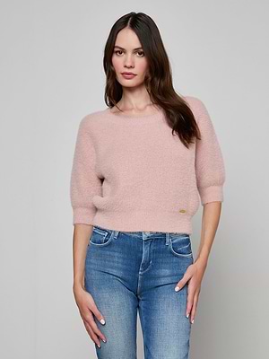 A woman stands against a plain background wearing the Guinevere Cropped Pullover, a light pink ultra-soft knit crewneck sweater with three-quarter sleeves, paired with blue jeans, looking at the camera with a neutral expression.