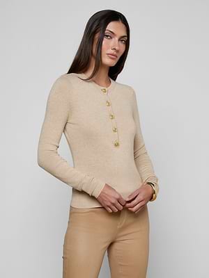 A woman with long brown hair models the Ronda Knit Henley Top in beige with gold button placket, paired with matching beige pants, standing against a light gray background.