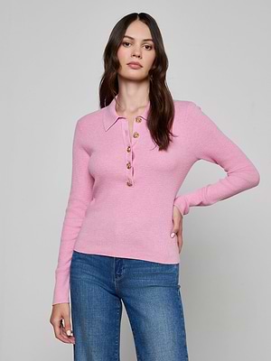 A woman with long brown hair and a neutral expression poses against a plain background, wearing the Sterling Sweater—a slim fit, collared pink knit with gold buttons—paired with blue jeans.
