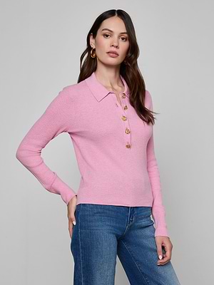 A woman with long brown hair and gold hoop earrings stands against a plain background, wearing the light pink Sterling Sweater—a slim fit, long sleeve knit henley top with gold buttons—paired with blue jeans.